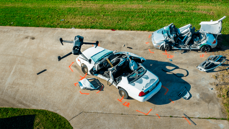 Дрон DJI Enterprise проводит съёмку места автомобильной аварии для реконструкции. На площадке видны два повреждённых автомобиля с разметкой и демонтированными деталями, подготовленными для анализа происшествия
