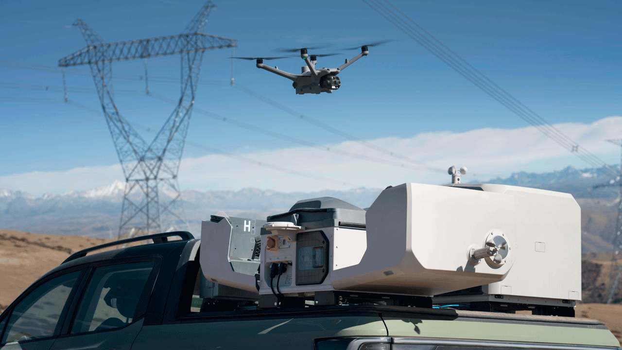 Дрон DJI Matrice в полете рядом с линиями электропередач, выполняющий инспекцию. На переднем плане автомобиль с установленной на крыше док-станцией DJI Dock 3. На заднем плане видны горы и голубое небо
