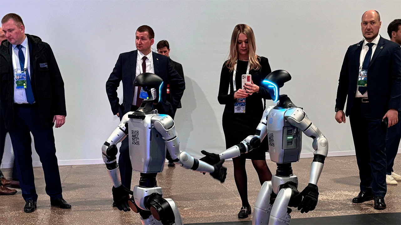 Два робота-гуманоида G1 демонстрируют взаимодействие друг с другом на Digital Almaty 2025, вокруг стоят посетители в деловой одежде с бейджами форума, наблюдая за презентацией