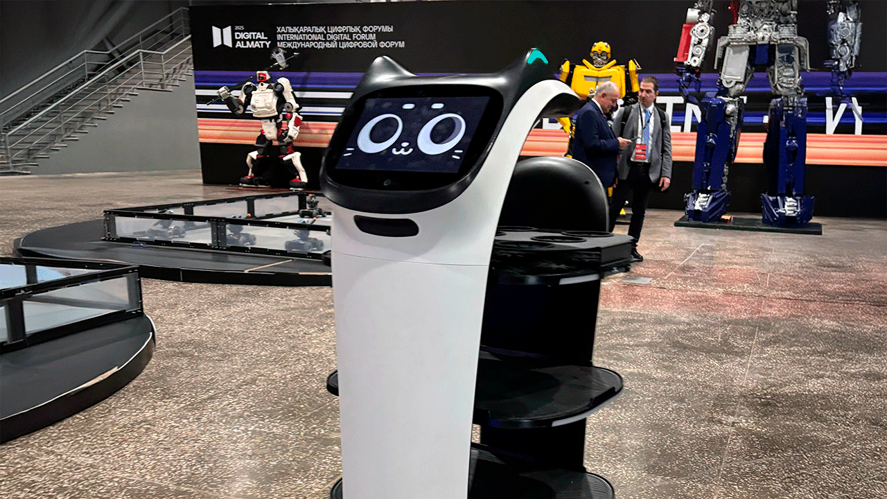 Робот-официант BellaBot от Pudu Robotics с милым анимированным дисплеем в виде кошачьей мордочки на выставке Digital Almaty 2025, на заднем плане видны другие роботы и баннер форума