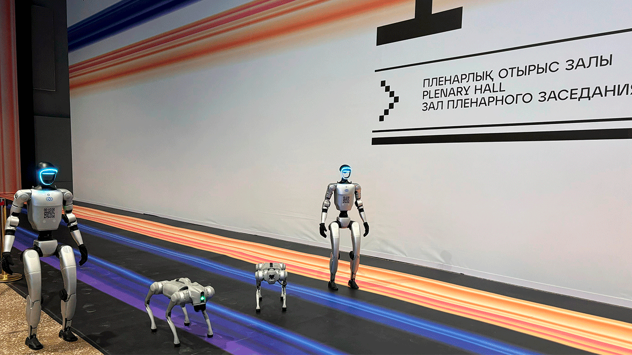Роботы Robot.kz у входа в пленарный зал Digital Almaty 2025: два робота-гуманоида G1 и два робота-пса Go2 демонстрируют свои возможности на фоне стены с официальной навигацией форума на трех языках