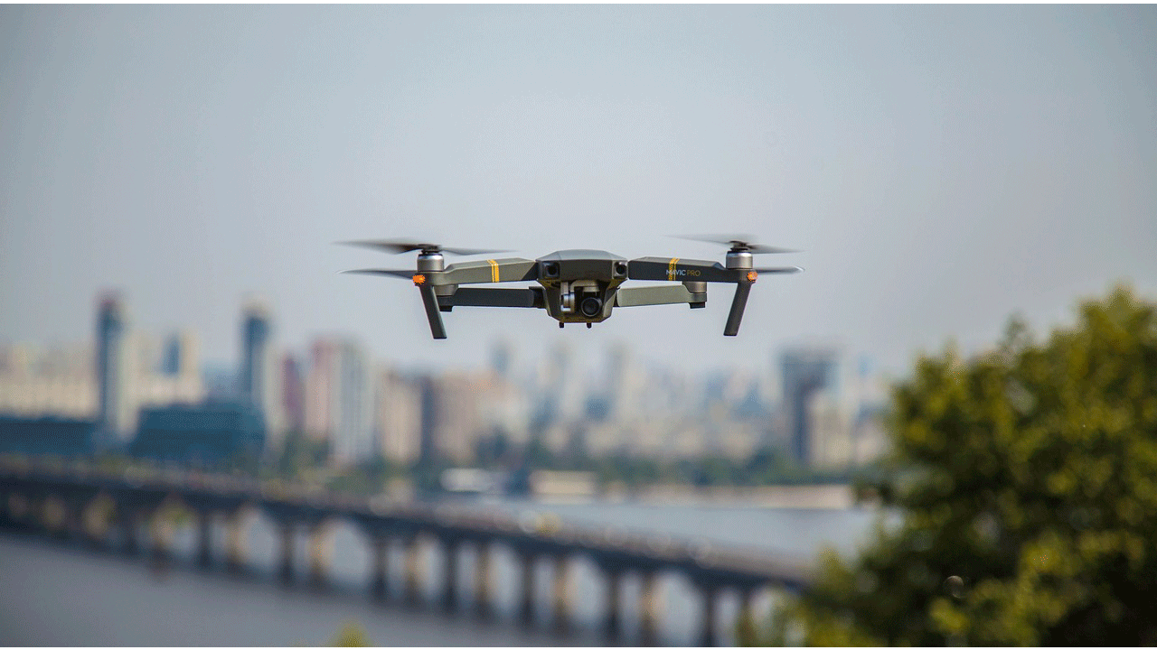 Квадрокоптер DJI Mavic в полете над городом, с размытым городским пейзажем и мостом на заднем плане