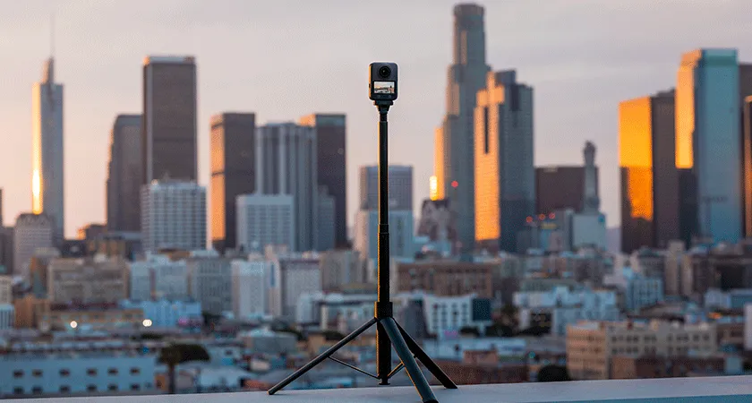 Экшн-камера DJI Osmo 360 на штативе с невидимой селфи-палкой на фоне городской панорамы при закате — демонстрация 360°-съёмки в урбанистической среде